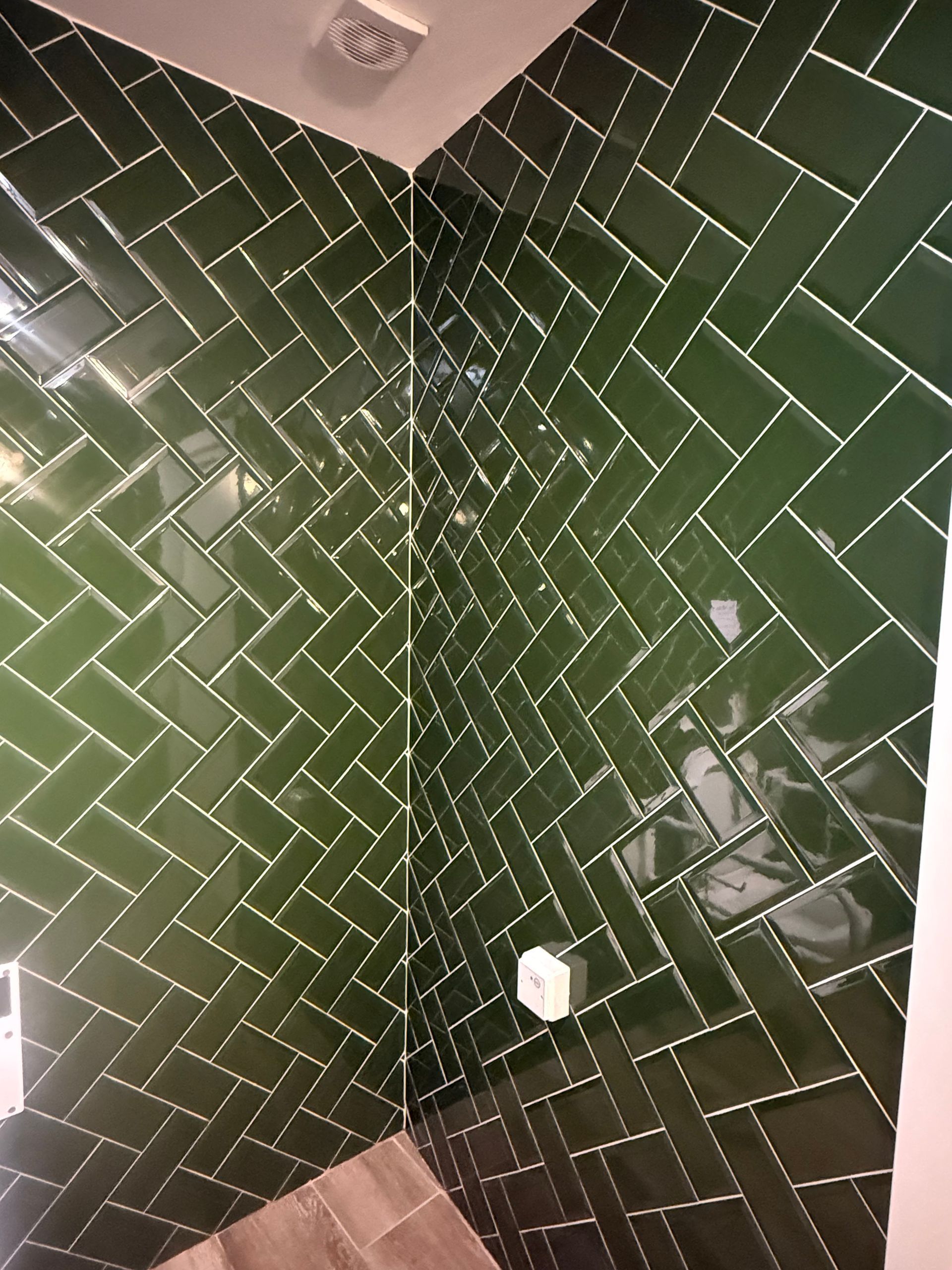 A corner shower wall featuring glossy, dark green subway tiles arranged in a herringbone pattern with white grout.