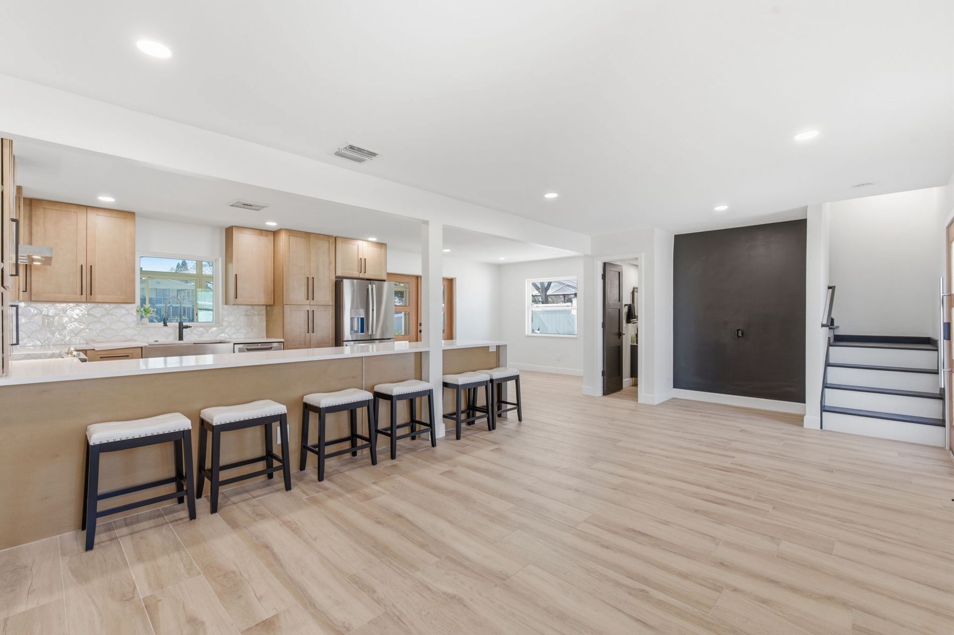 A modern, open-concept kitchen with light wood cabinets, a breakfast bar with five stools, and light wood floors.