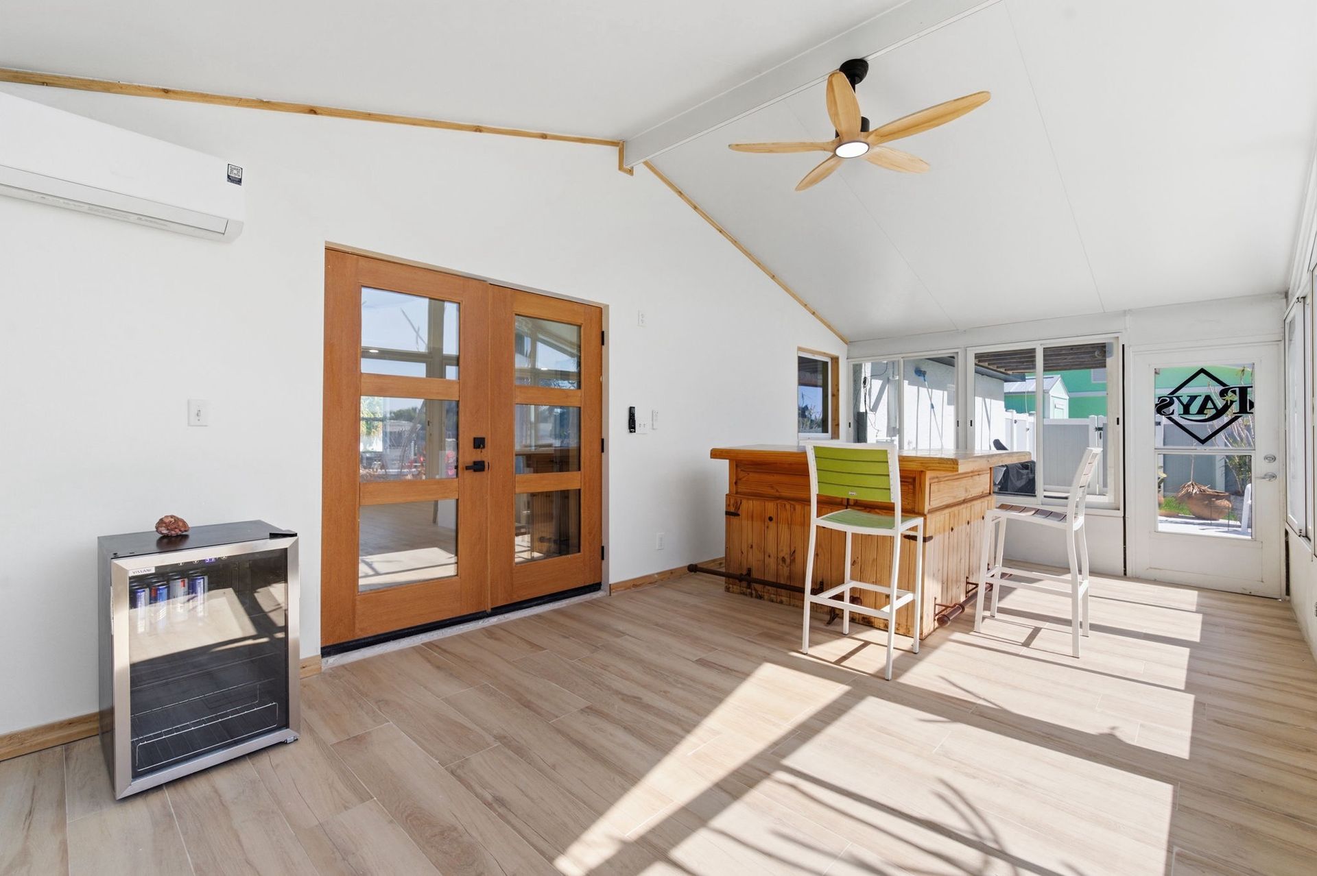 A bright sunroom with light wood flooring, a beverage cooler, a wooden bar with two high stools, and a ceiling fan.