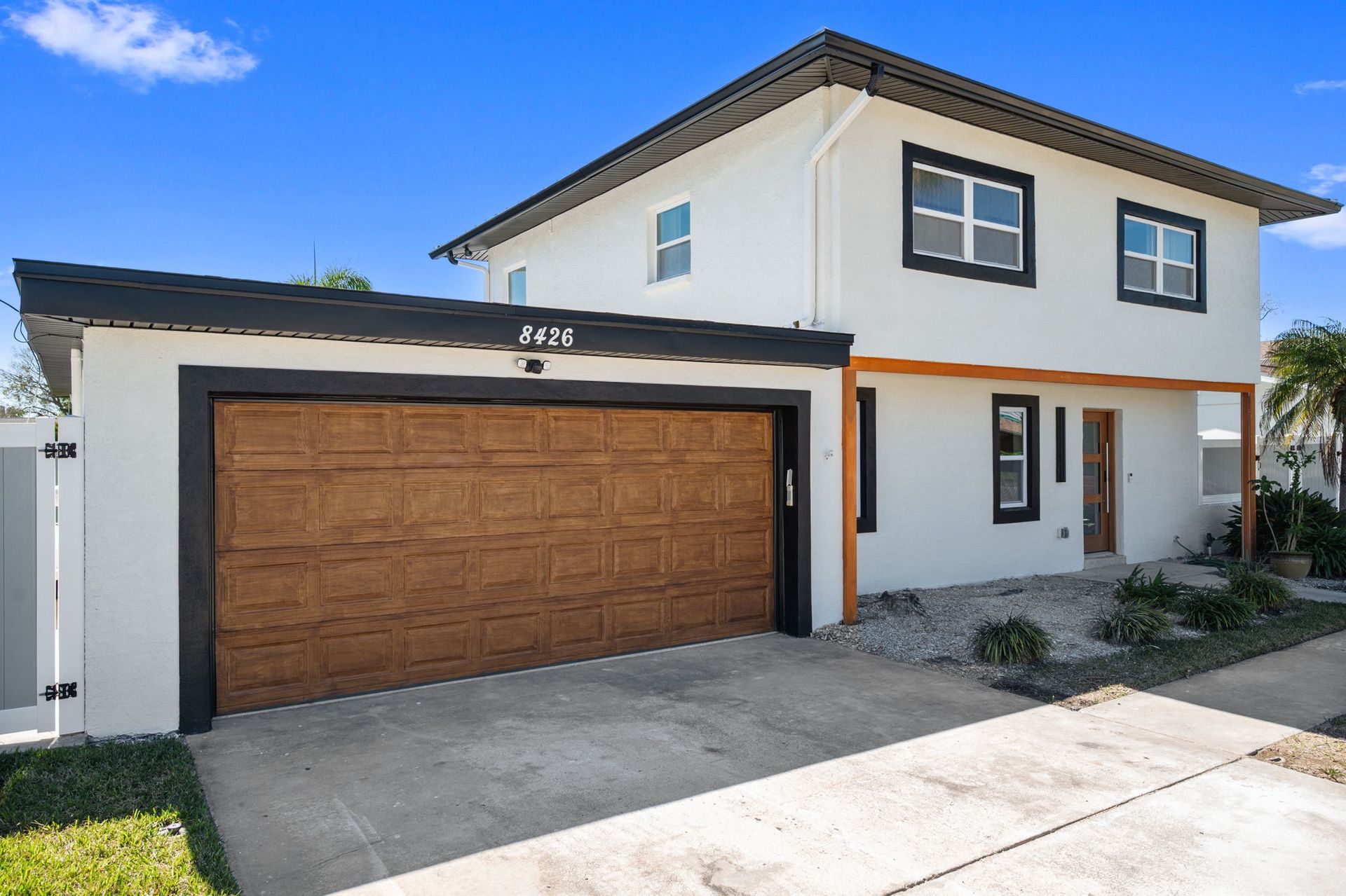 A white two-story house with a wooden garage door, black trim, and a concrete driveway under a bright blue sky.