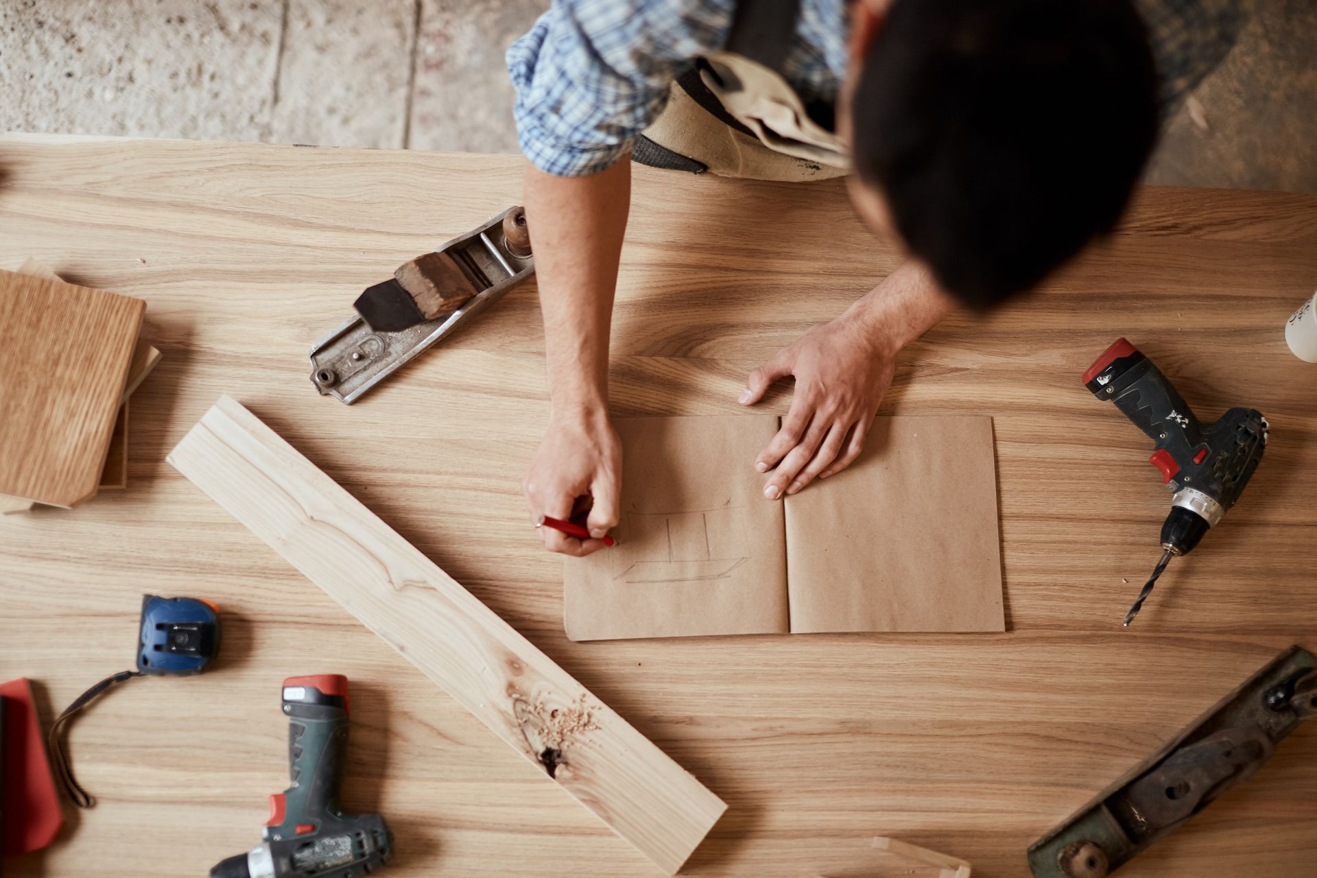 Carpenter sketches design on paper surrounded by tools on a wooden workbench.