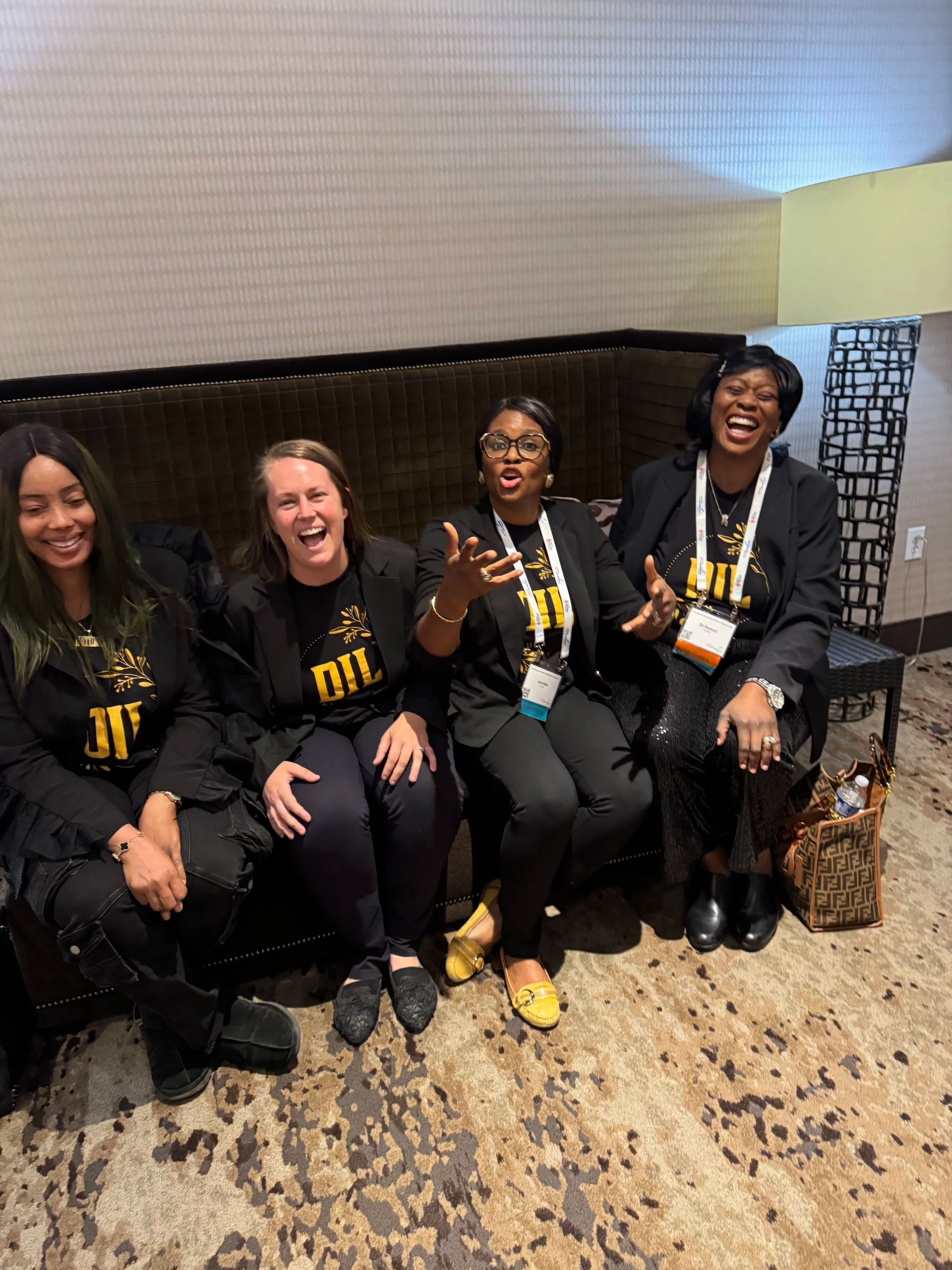 Four smiling people in black shirts sit on a bench indoors, wearing lanyards and holding cups.