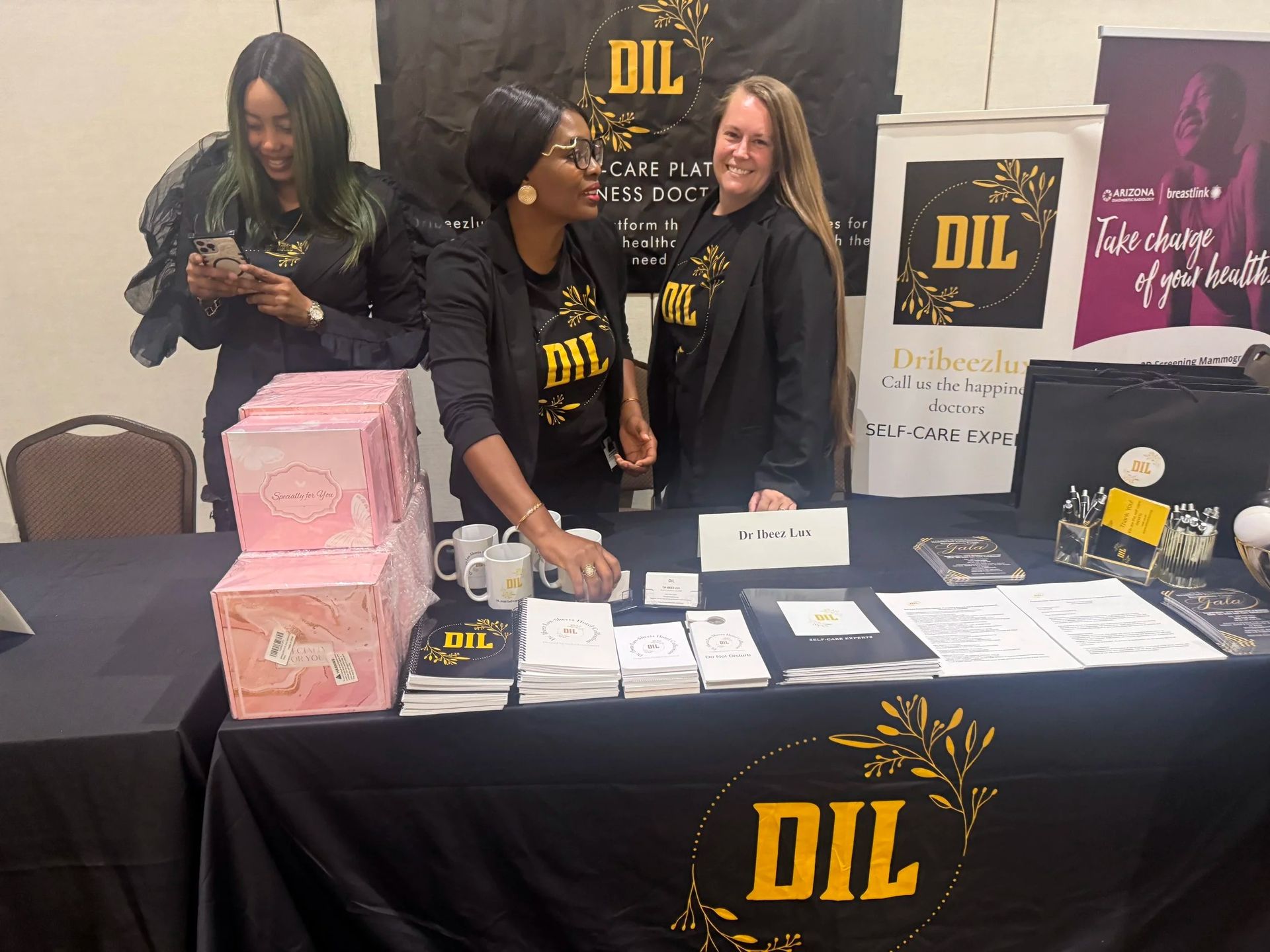 Three people at a DIL event booth with black tablecloth, flyers, brochures, and pink stacked boxes