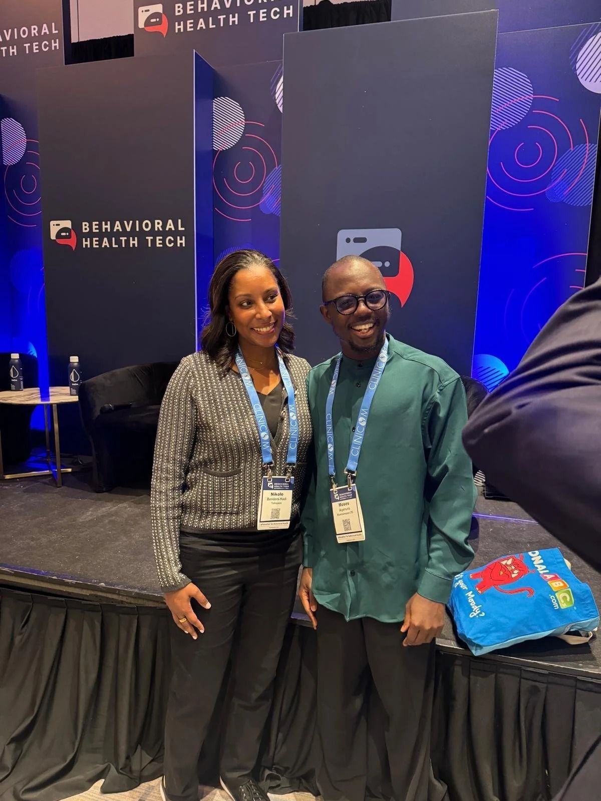 Two smiling people pose at a conference booth with a black-and-blue backdrop and event lanyards.