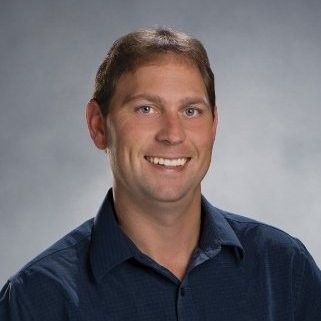 Man with short brown hair smiling, wearing a blue button-down shirt, studio shot.