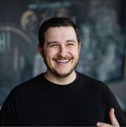 Man with dark hair and beard smiling broadly in front of a blurry background. He wears a black shirt.