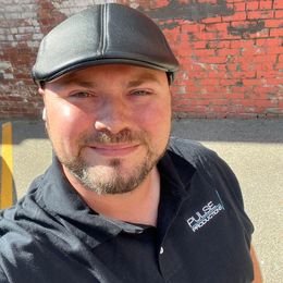 Man wearing a black hat and shirt smiles in front of a brick wall.