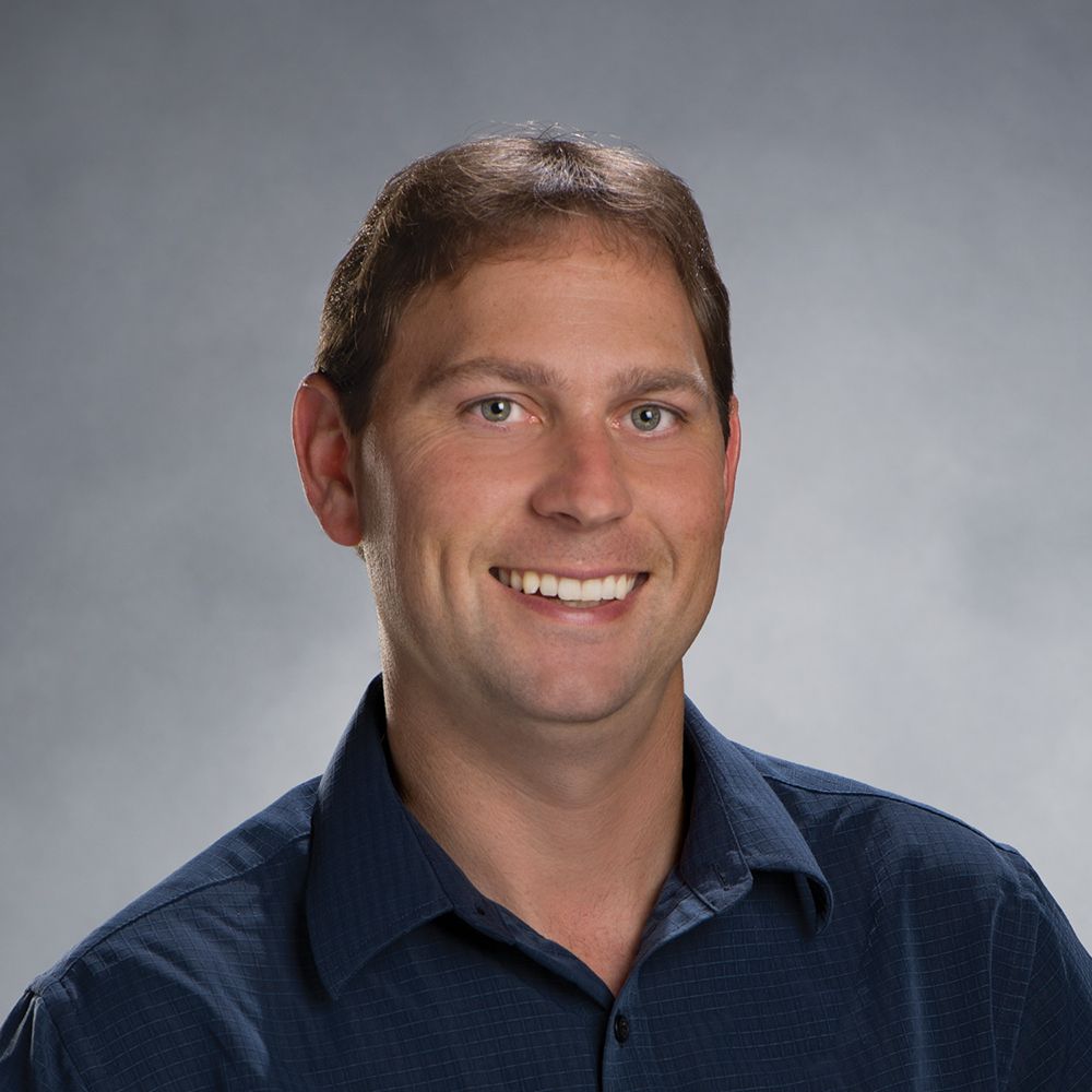 Man with short brown hair smiling, wearing a blue button-down shirt, studio shot.
