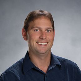 Man with short brown hair smiling, wearing a blue button-down shirt, studio shot.