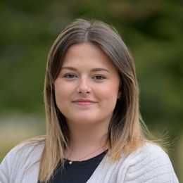 Woman with blonde hair, smiling, wearing a black shirt and white cardigan. Outdoors with green background.