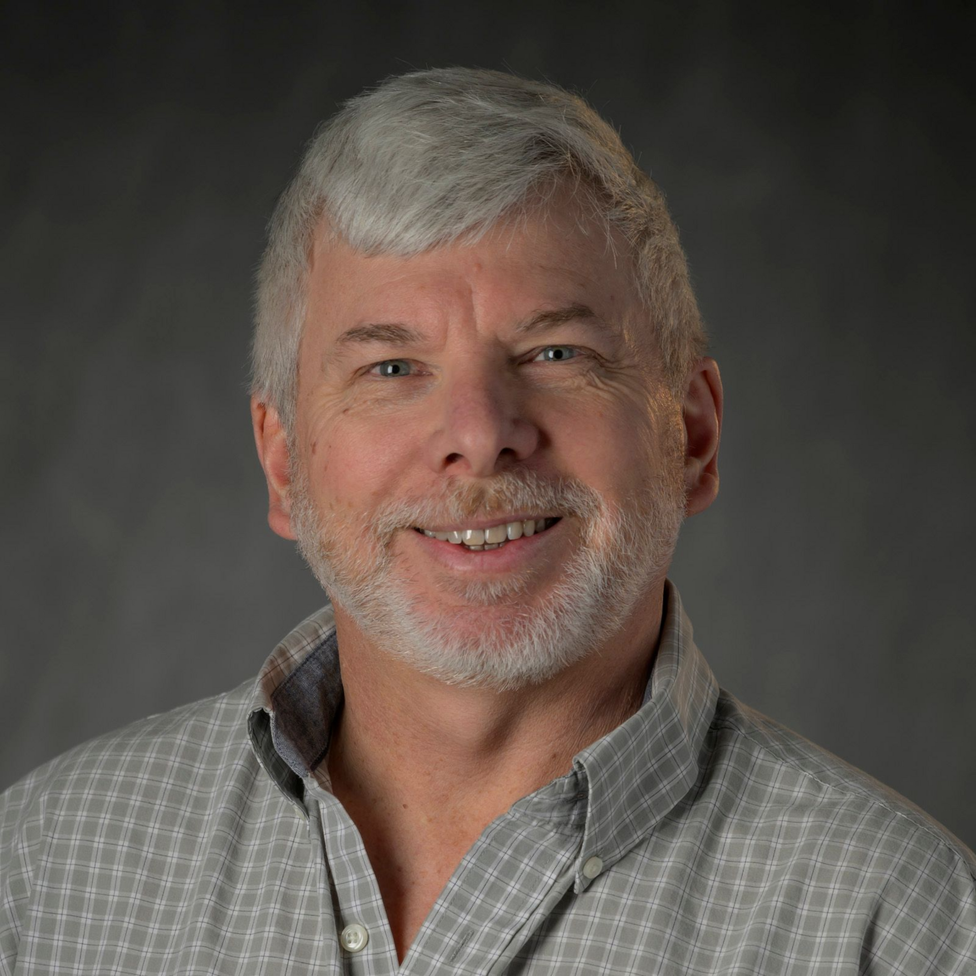 Woman with blonde hair, smiling, wearing a black shirt and white cardigan. Outdoors with green background.
