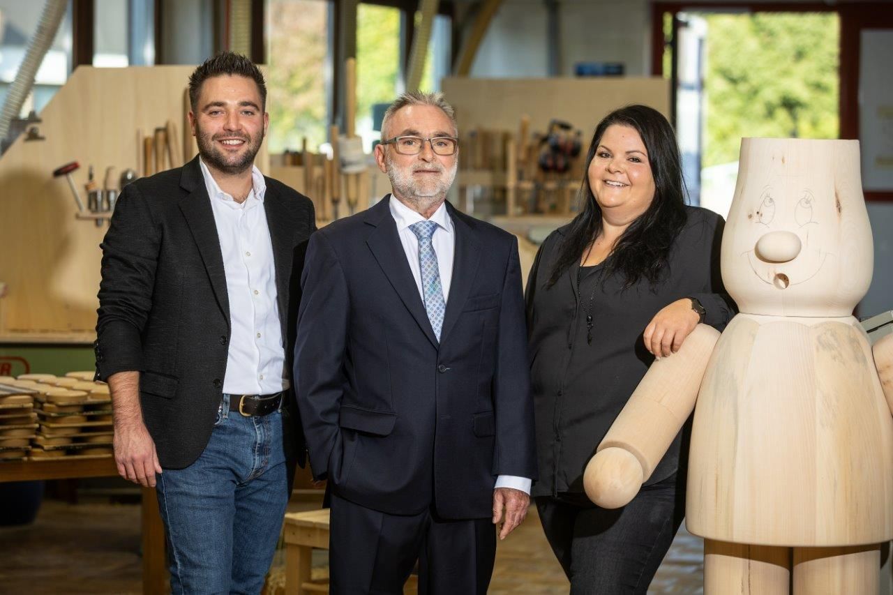 Three people in a workshop pose with a large wooden doll.