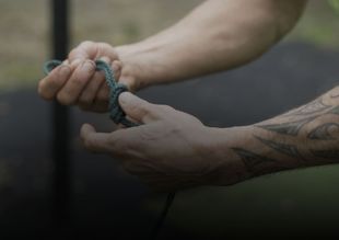 Handen die buiten een touw vastknopen, waarbij een getatoeëerde onderarm zichtbaar is.