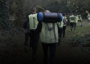 Een groep wandelaars in reflecterende hesjes en rugzakken loopt over een donker, bebost pad.
