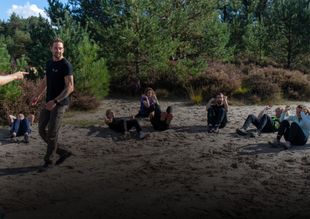 Mensen zitten en liggen op de zanderige grond in een open plek in het bos, met één persoon die vooraan staat.