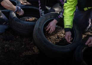 Mensen plaatsen banden rond planten in een bloembed, waarbij handen en een neonkleurige jas zichtbaar zijn.