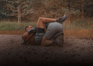 Twee mannen worstelen op een stoffig veld, waarbij de ene de andere vastklemt te midden van droog gras en bomen.
