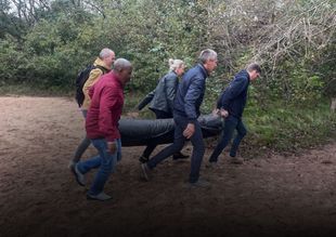 Vijf mensen dragen een grote band over een onverhard pad langs bomen en struiken.
