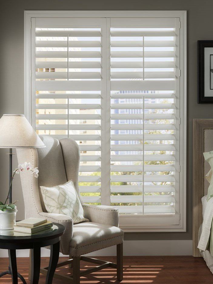 A living room with a chair , table , lamp and shutters on the window.