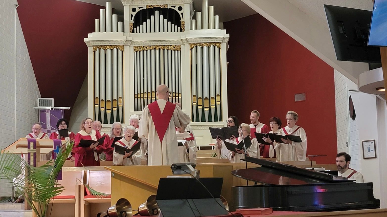 Church choir performing in front of a large pipe organ, with a piano in the foreground