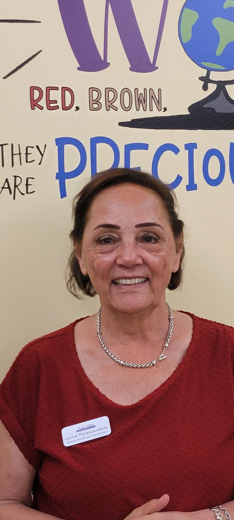 Smiling woman in red shirt standing before a classroom wall with colorful words and a globe poster.