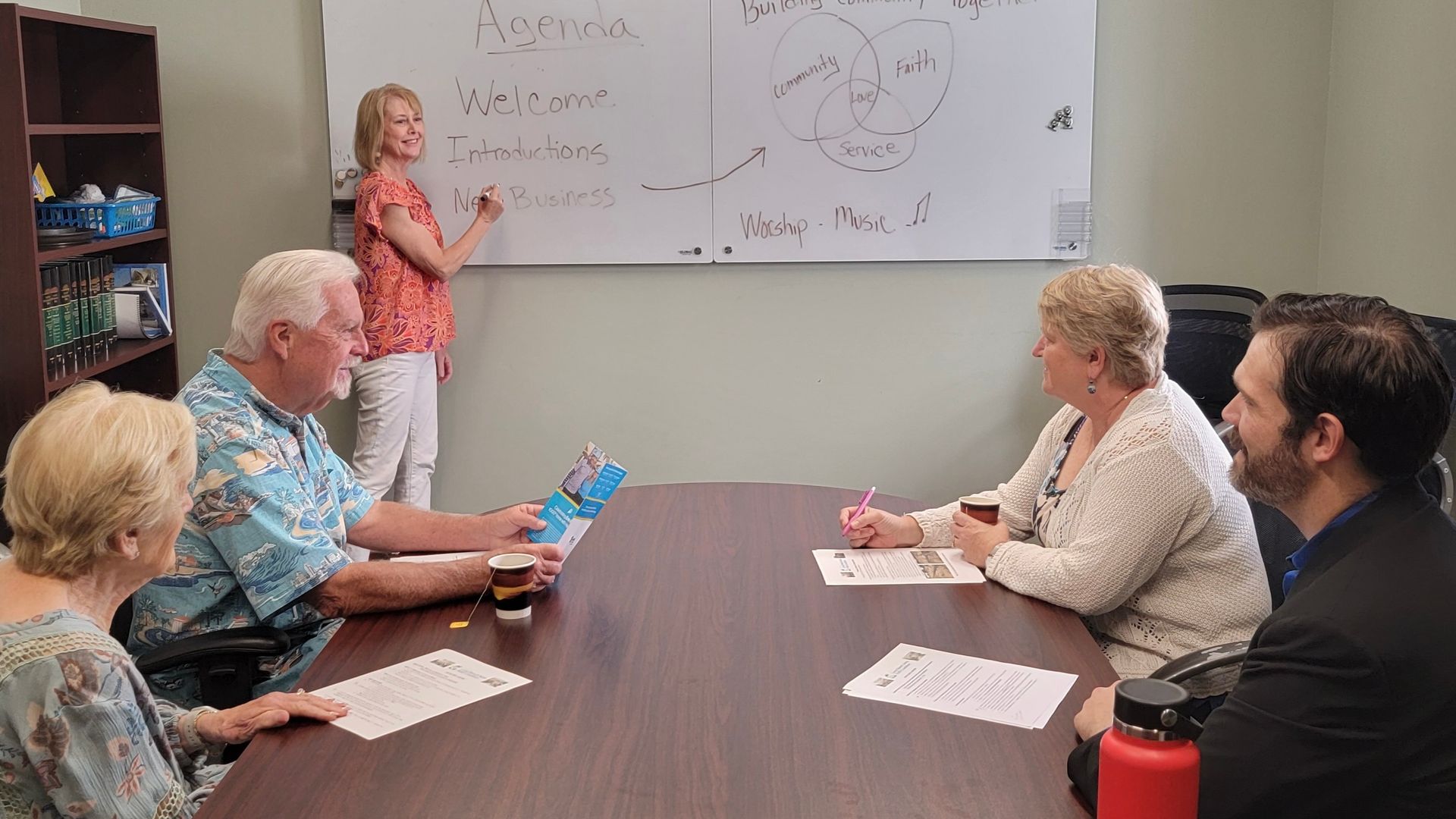 Small meeting room with a woman presenting at a whiteboard while four people sit around a conference table.
