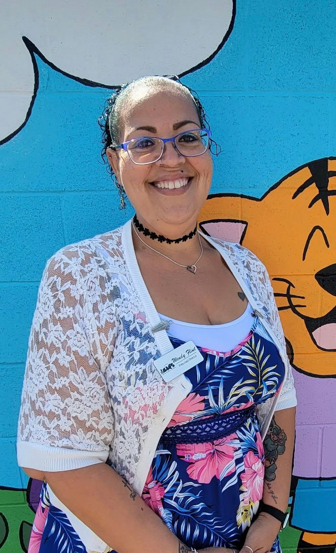 Smiling person in glasses standing before a colorful mural, wearing a white floral cardigan and patterned top