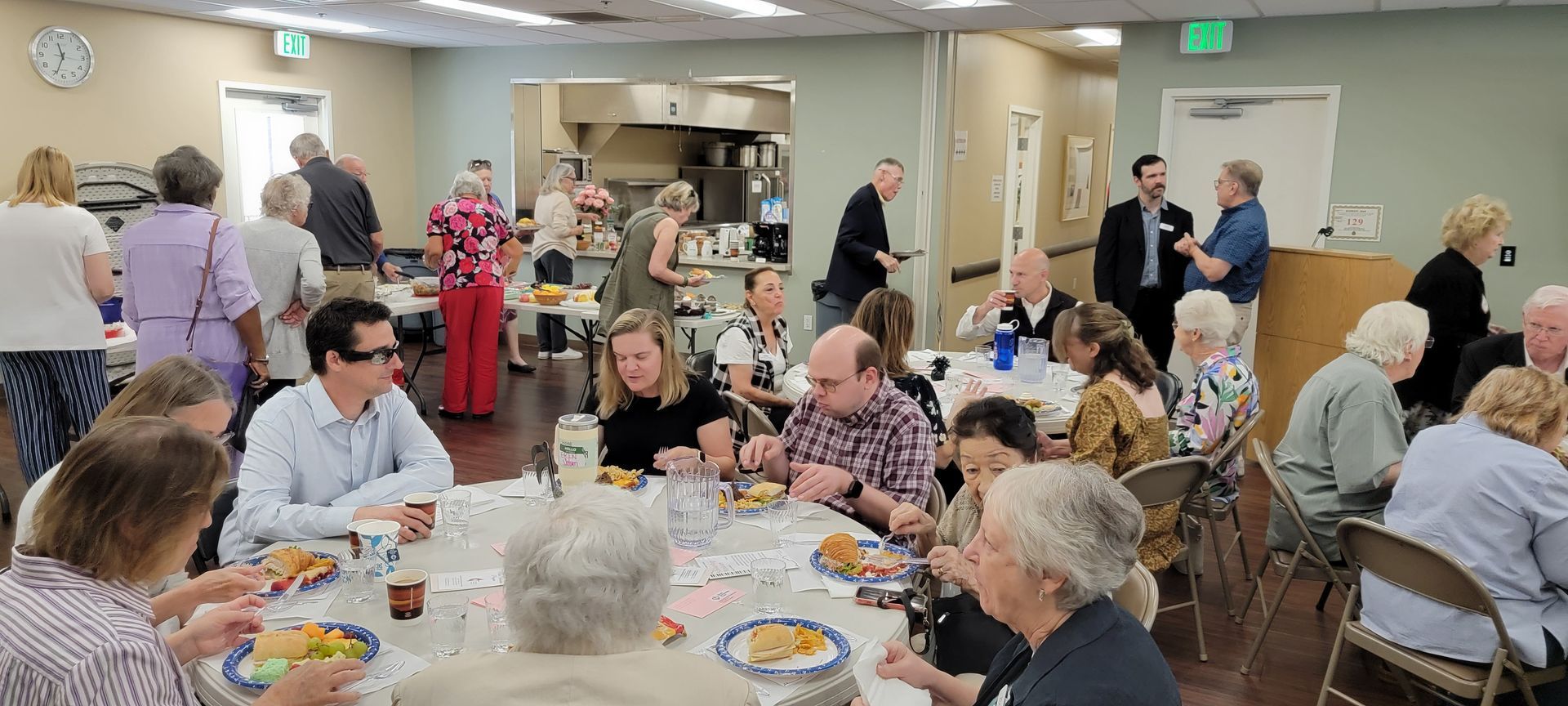 Community room with people seated at tables, eating and talking during a social gathering