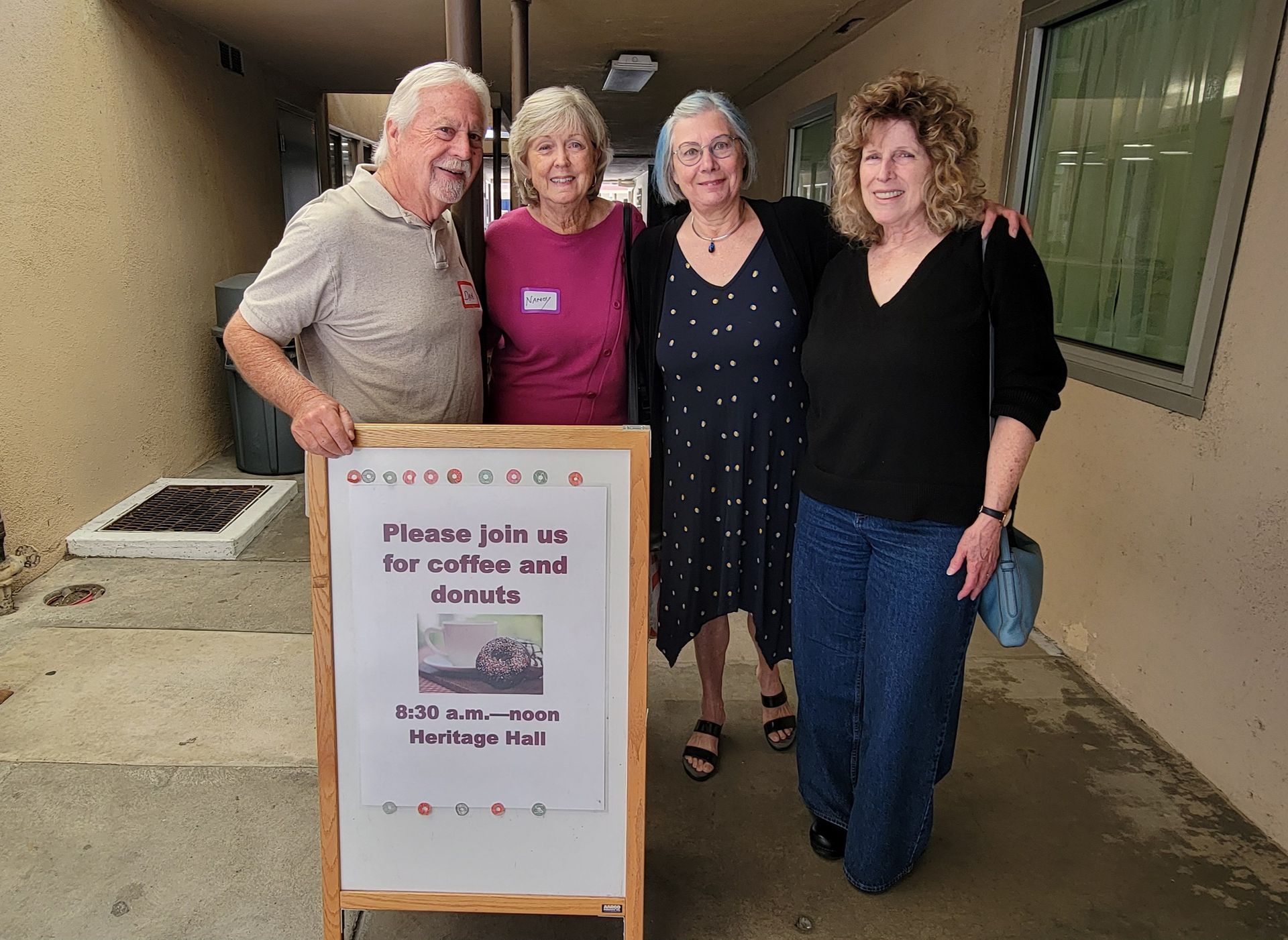Four adults pose beside a signboard outside a building, with a window and walkway in the background.