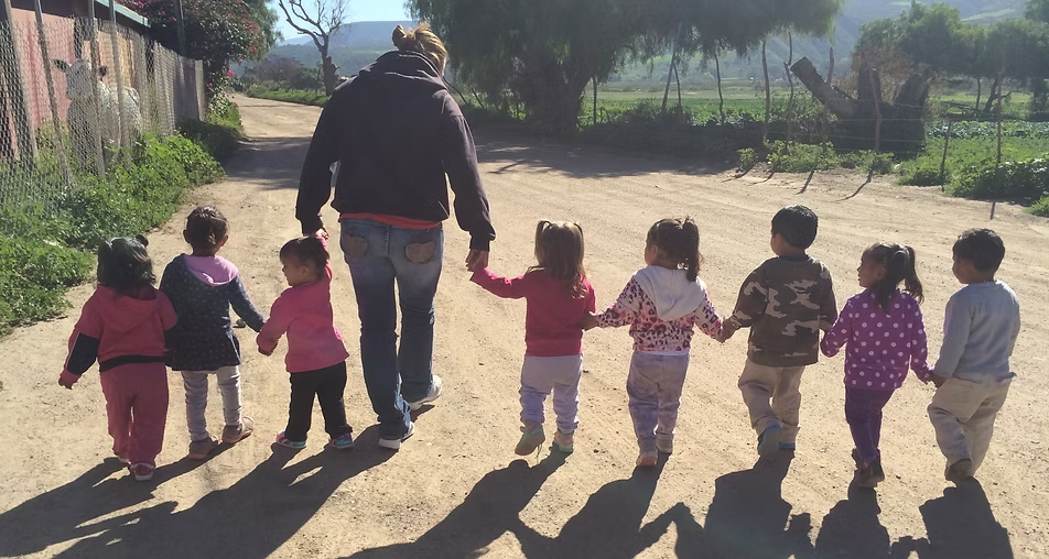 Adult holding hands with six children walking down a sunlit dirt path outdoors