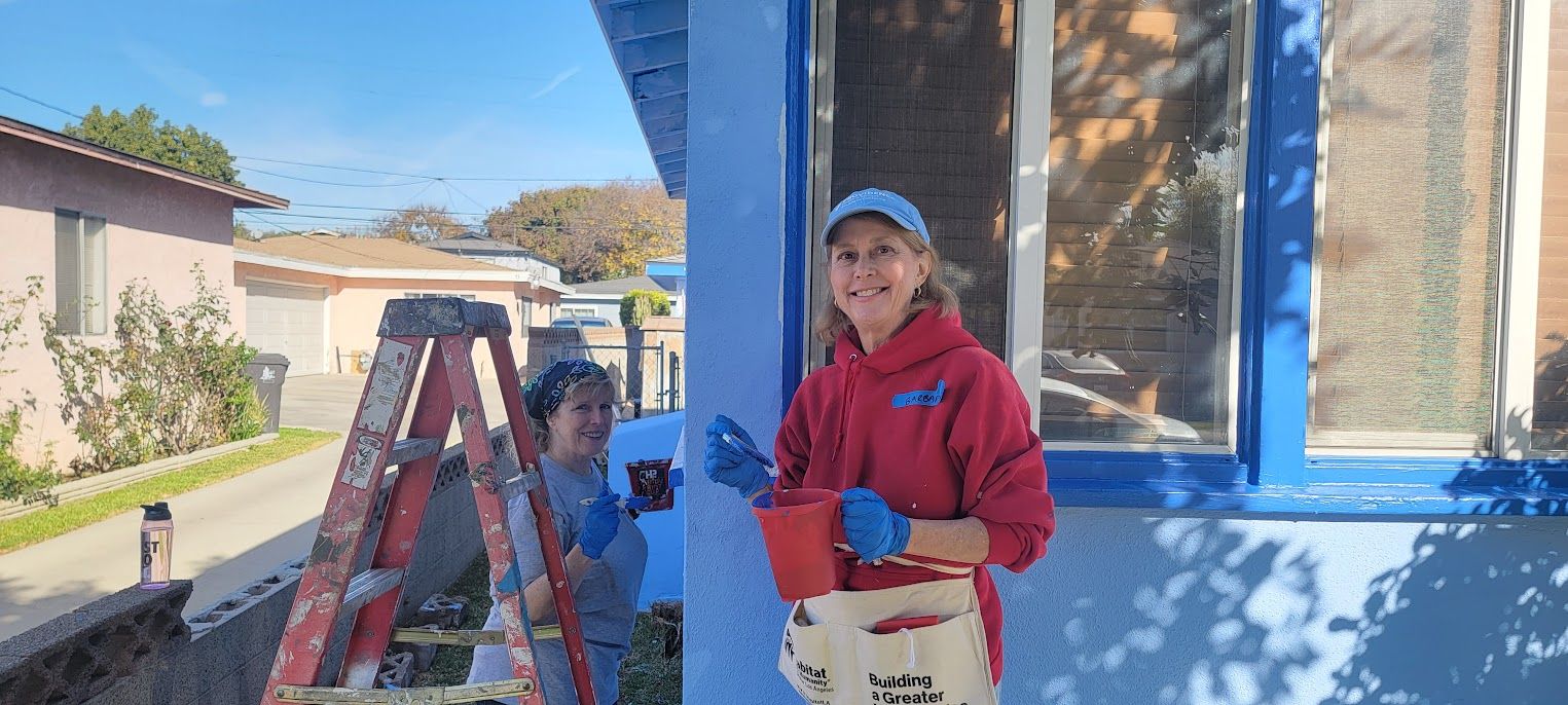 Person in red jacket and cap stands by a ladder painting a blue house exterior.
