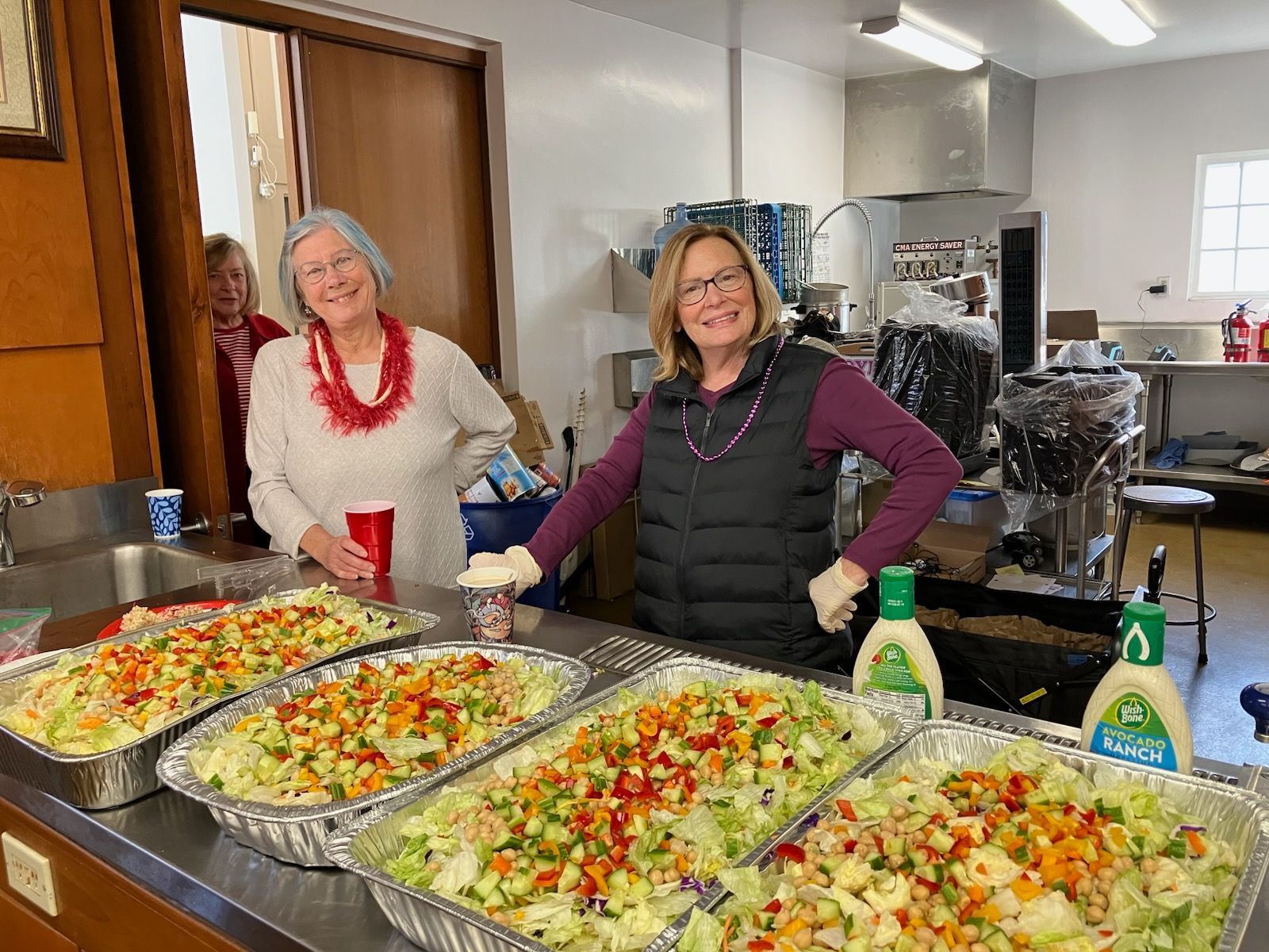 Two women serving colorful pasta salads at a buffet table in a kitchen.