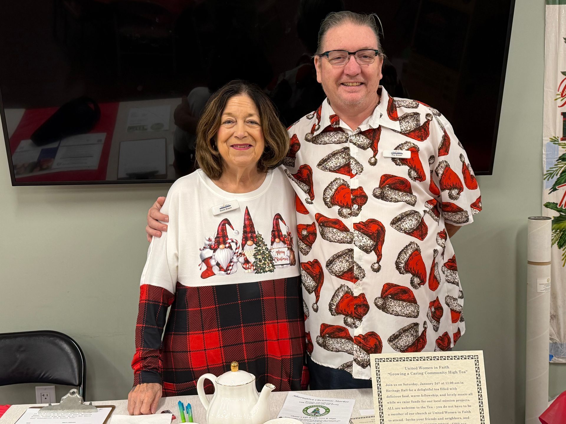 Two people standing behind a table, smiling in festive red-and-white holiday shirts