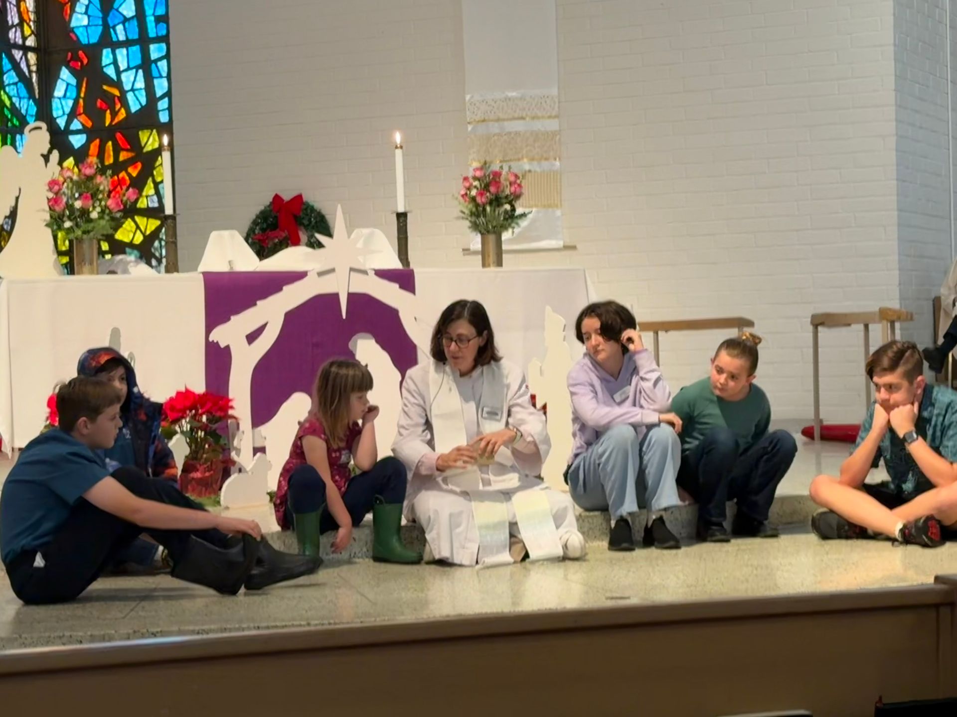 Children sit on a church stage near a purple nativity backdrop and candles.