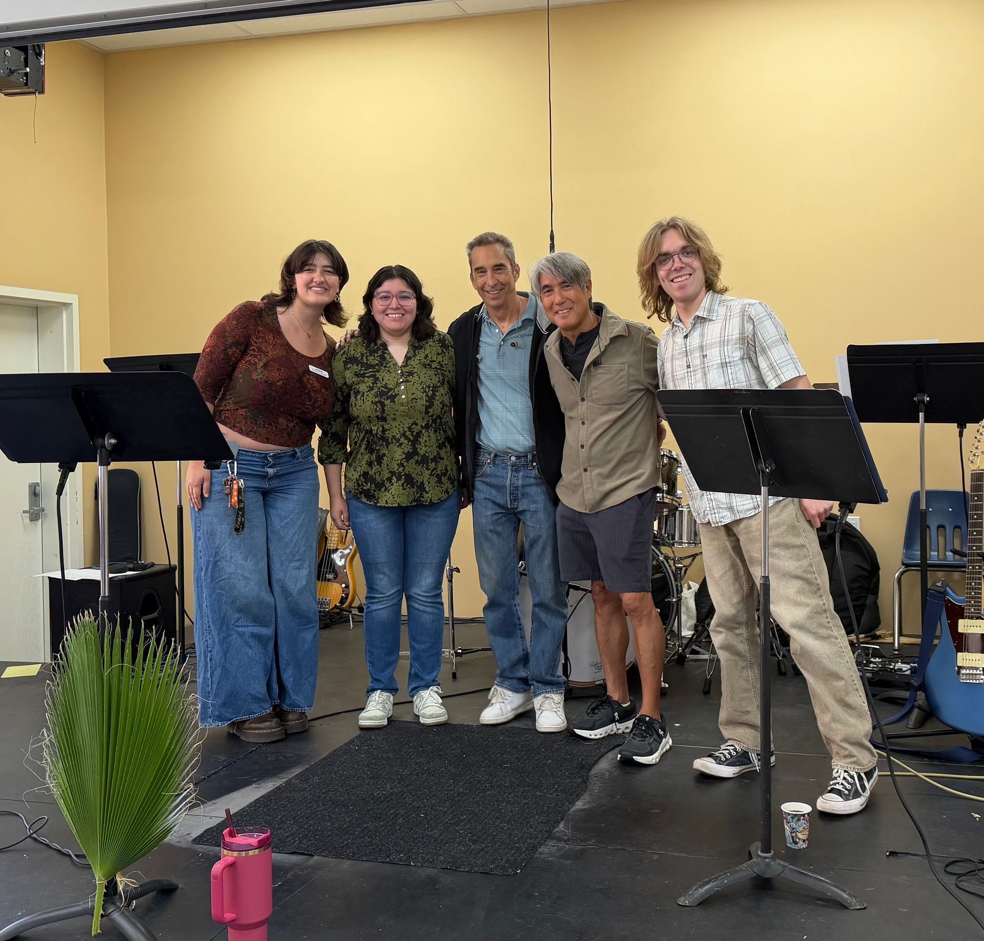 Five people pose smiling on a small stage with music stands and microphone stands.