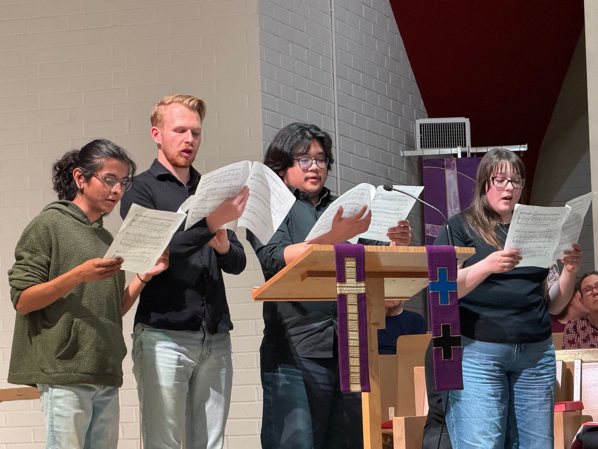 Four people stand behind a podium, holding sheet music and singing in a church.