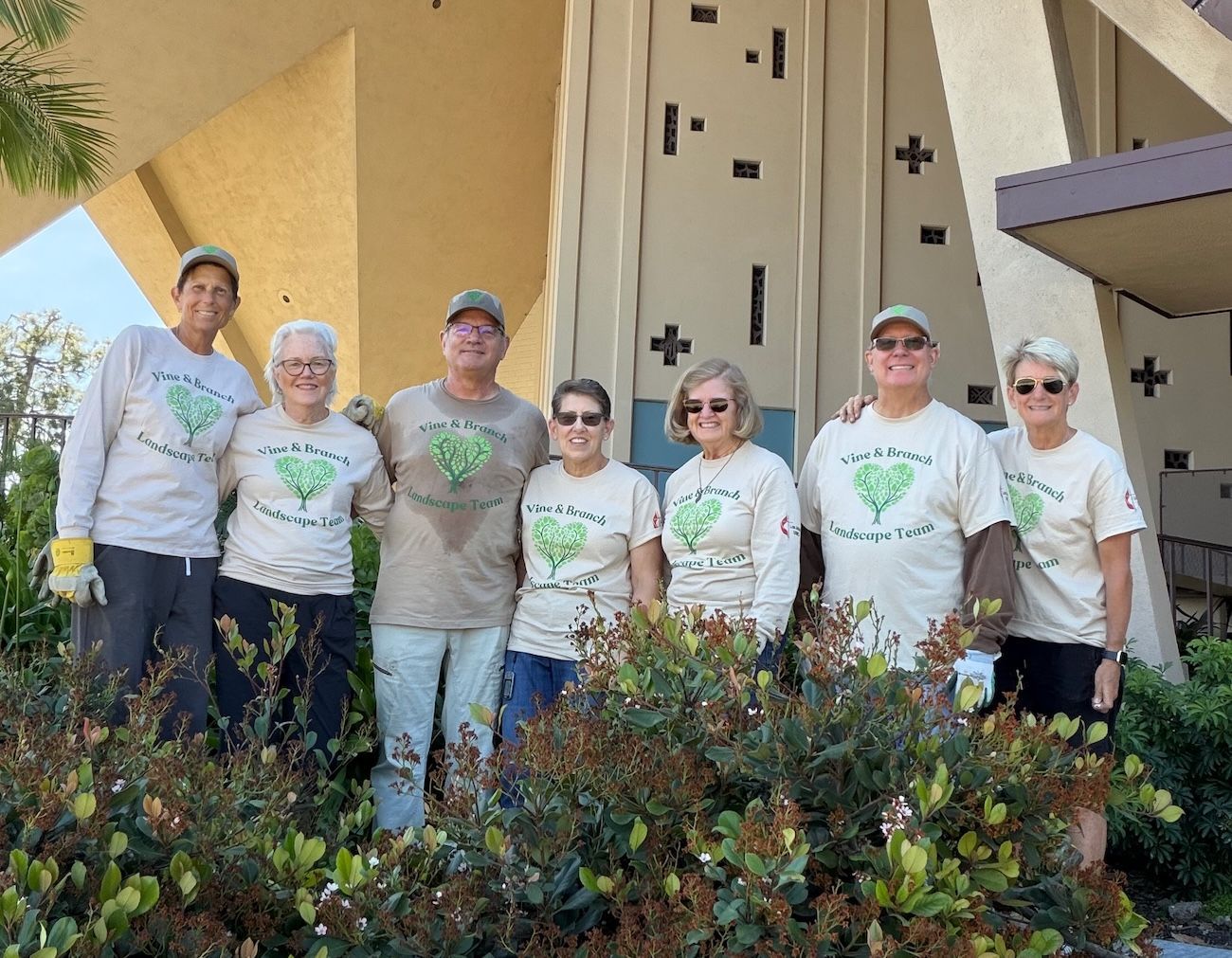 Seven volunteers in matching white T-shirts stand smiling outside a church among shrubs.