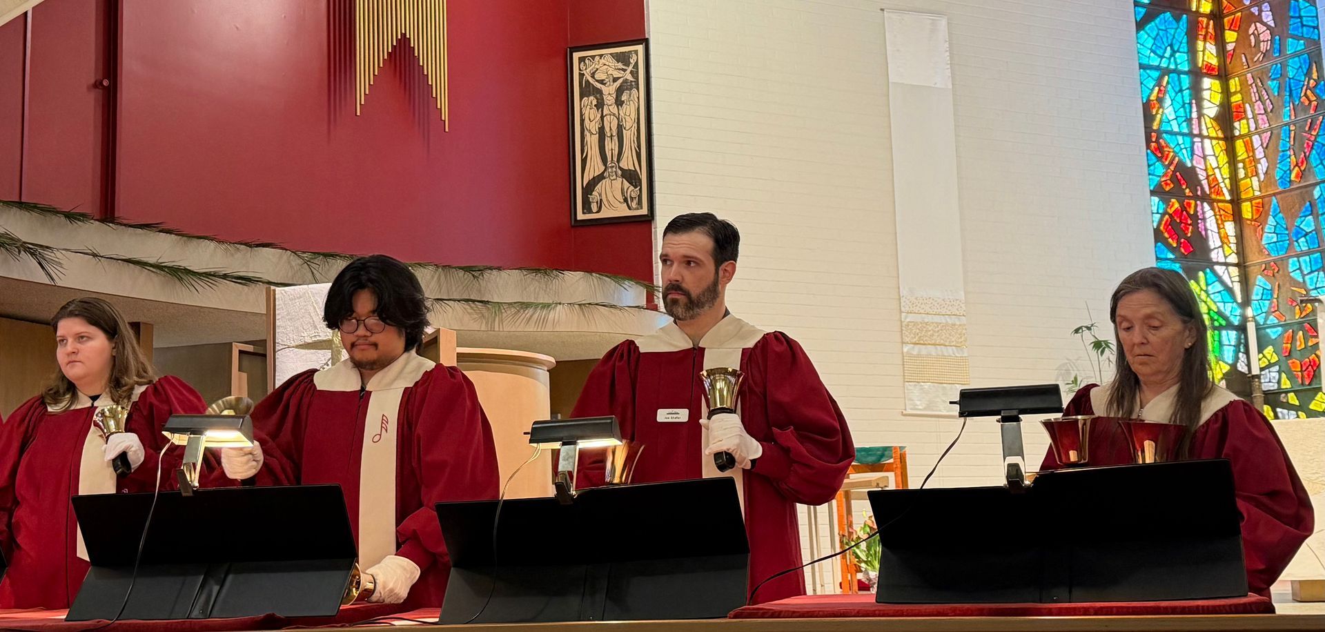 Persons in red robes playing bells at a church altar with stained glass behind them