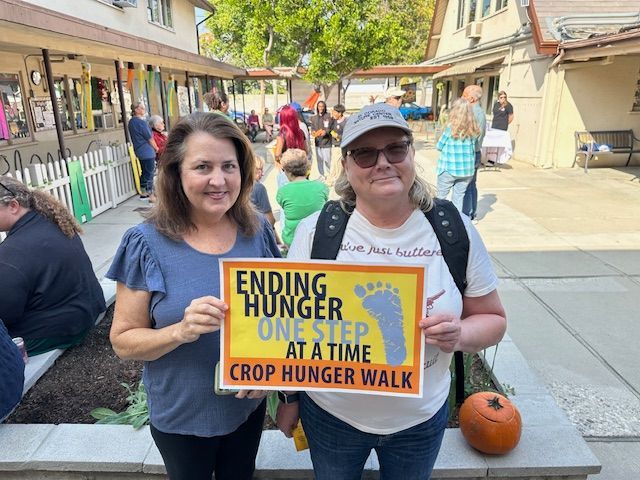 Two women holding an “Ending Hunger One Step at a Time” Crop Hunger Walk sign at an outdoor event