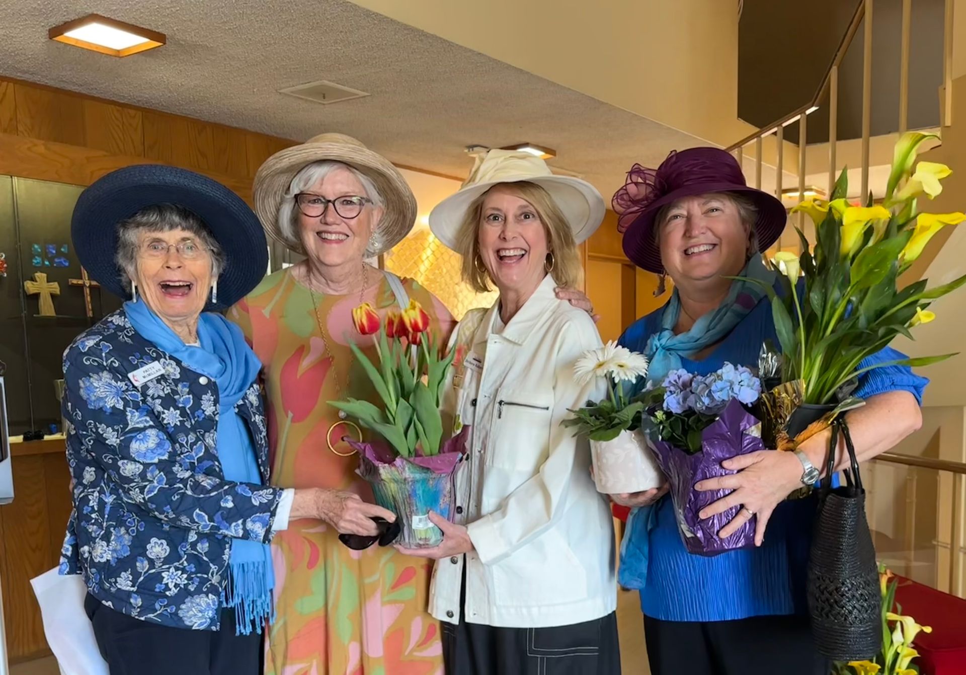 Five women in colorful hats hold bouquets indoors, smiling at a celebration.