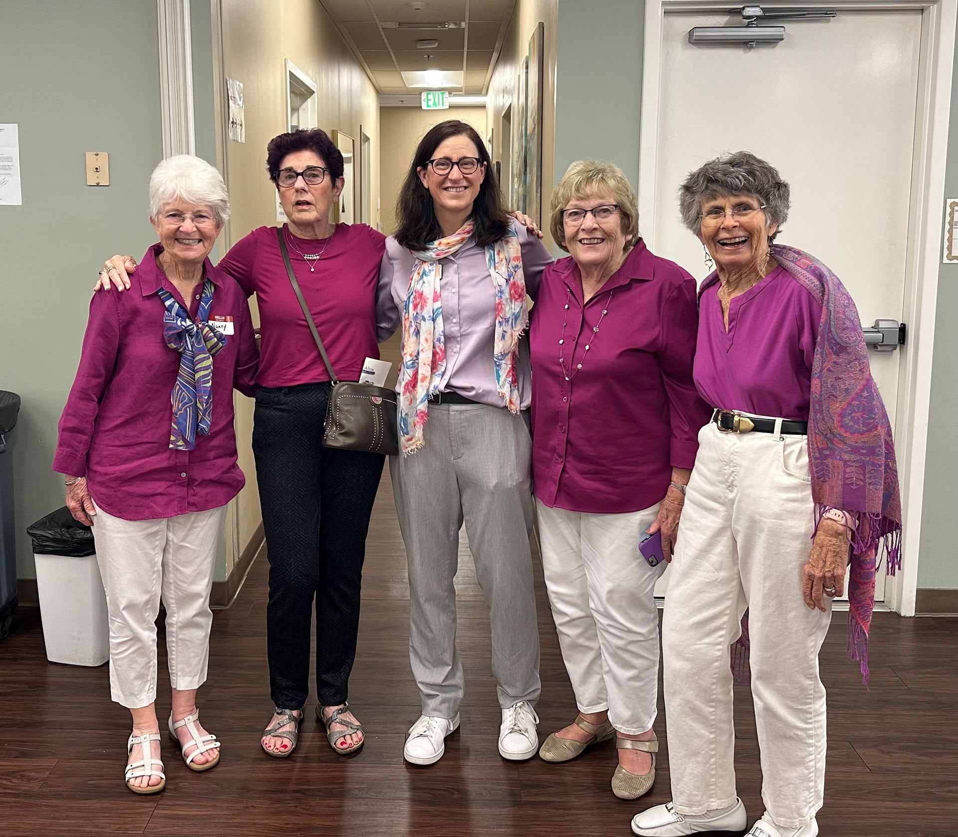 Five women in similar magenta tops pose smiling in a hallway.