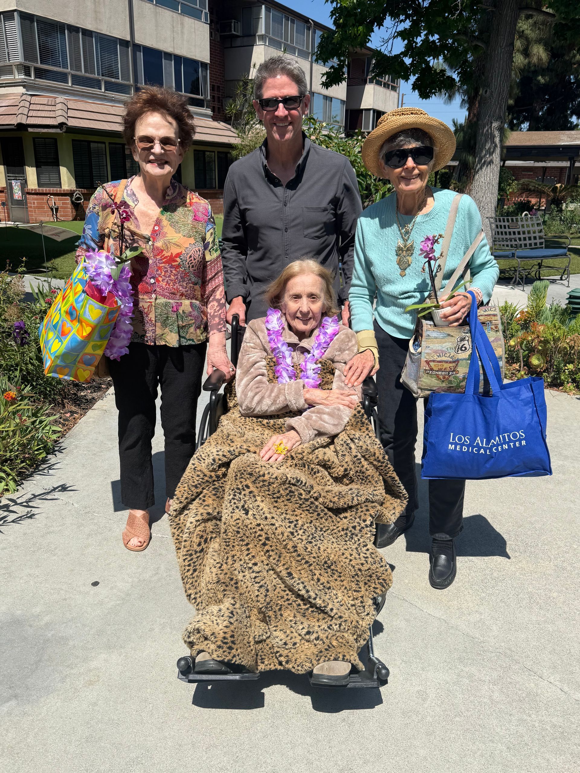 Four people smiling outdoors on a sunny walkway, one seated in a leopard-print blanket, others holding colorful bags.