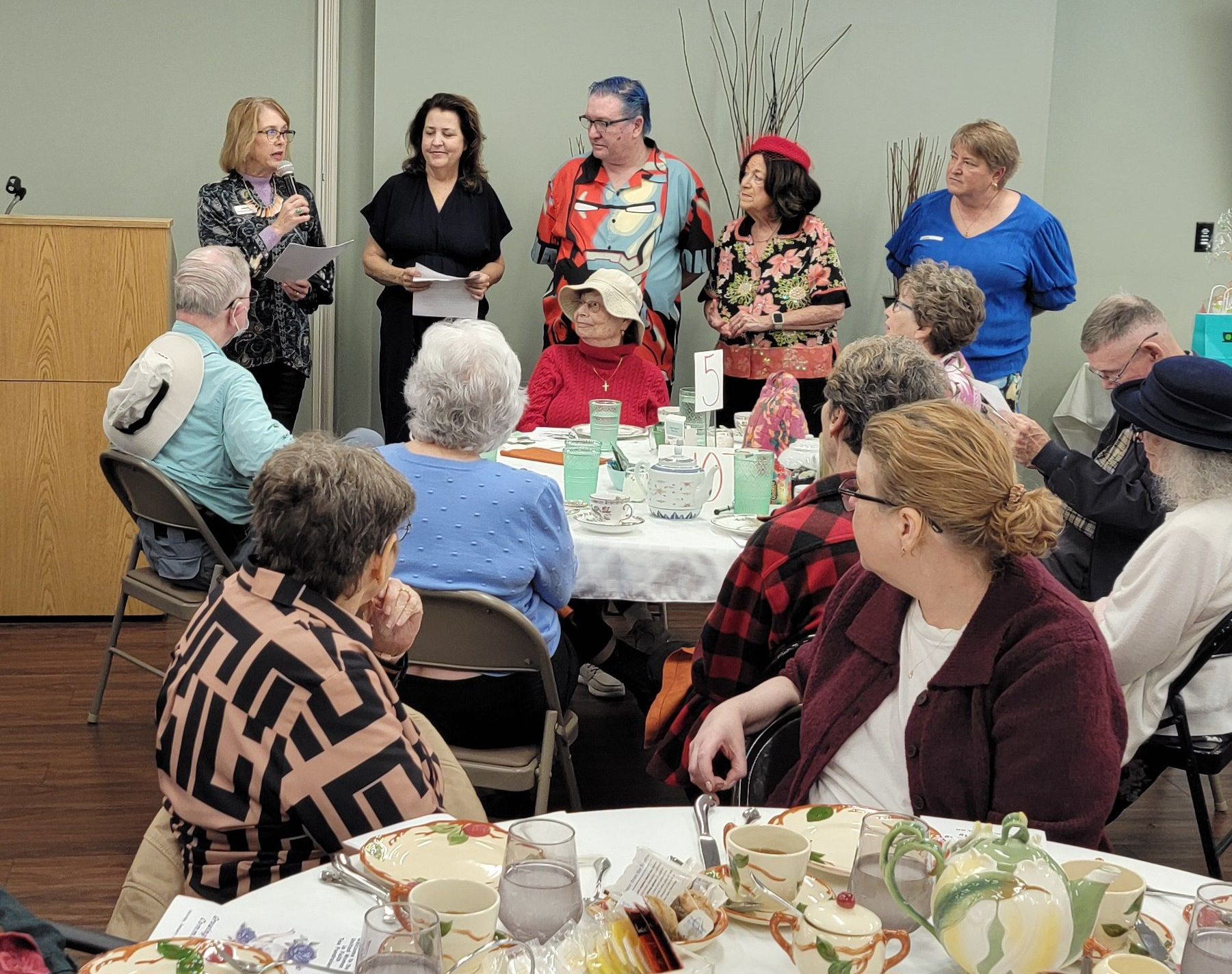 People gathered around tables in a bright room, listening to a speaker at the front.