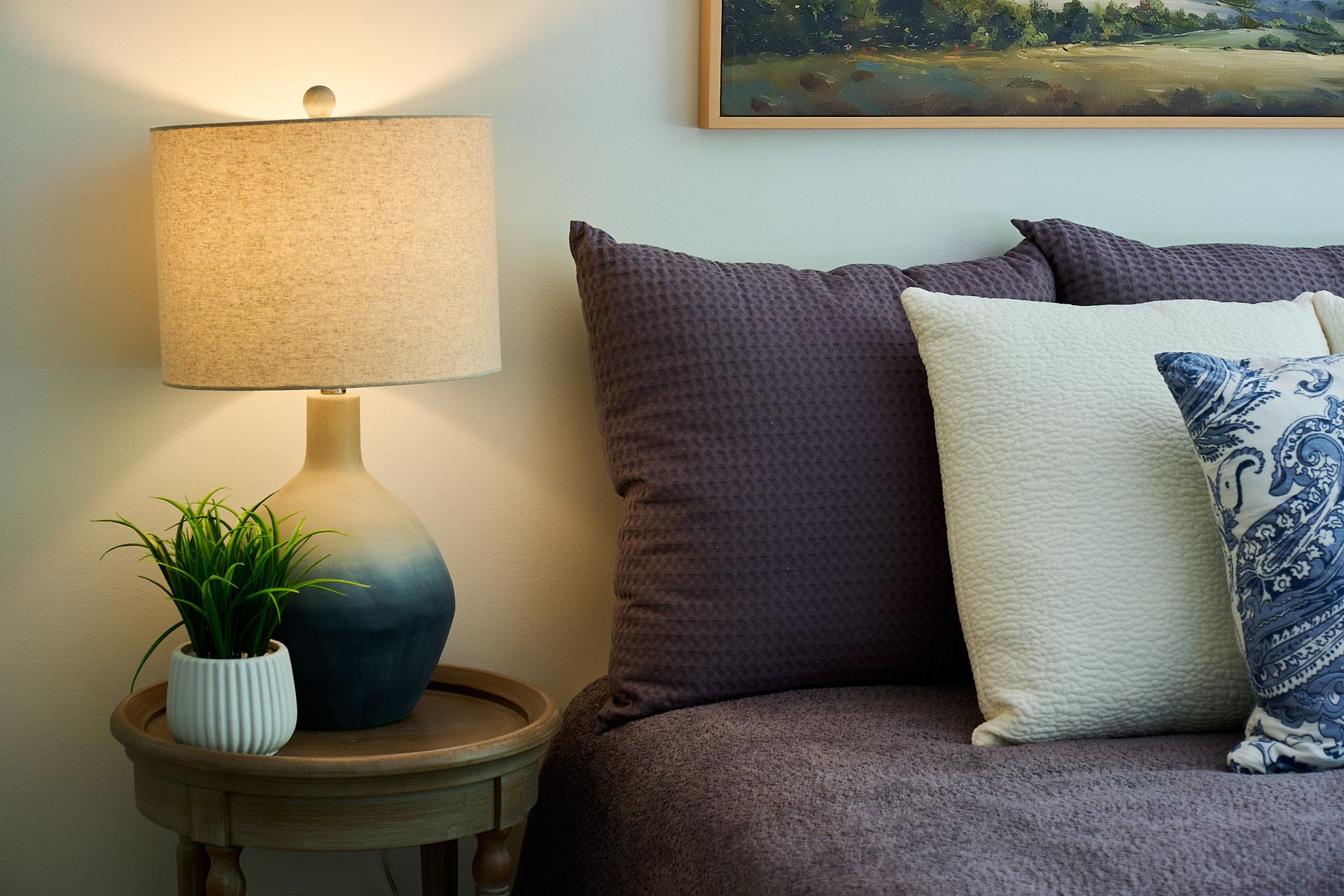 A bedside table with a lighted lamp and small potted plant, next to a bed with dark and white textured pillows.