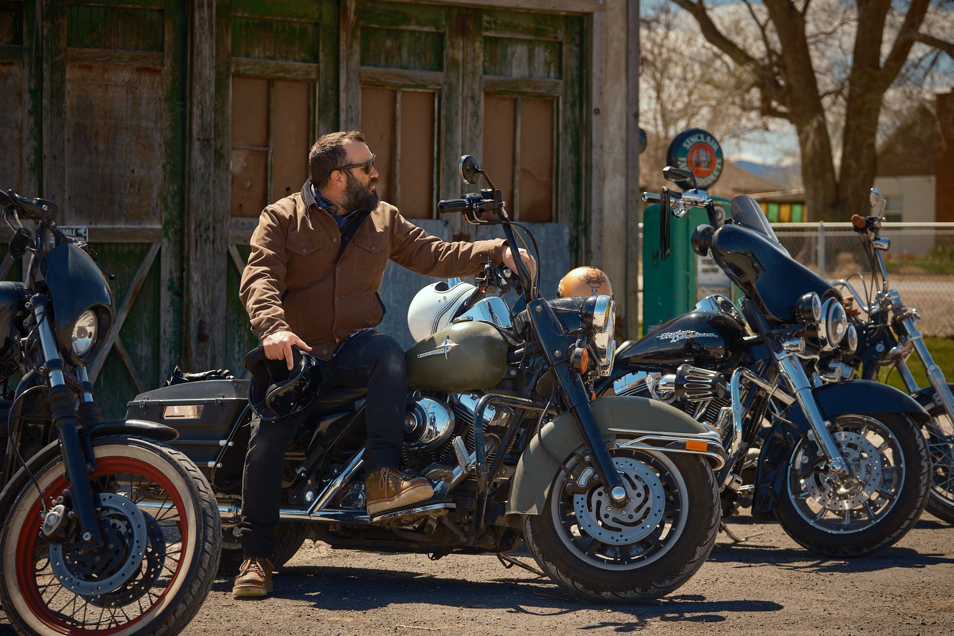 A person in a brown jacket sits on a motorcycle parked outdoors next to other motorcycles in front of an old garage.