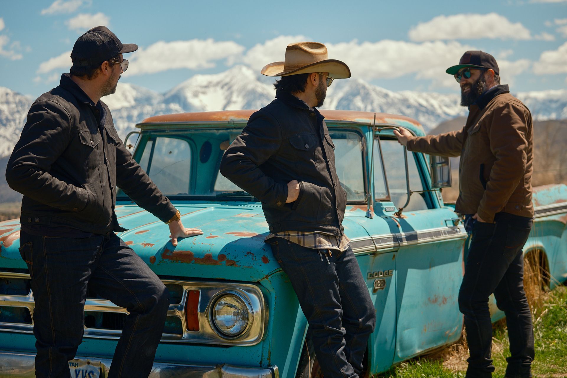 Three people stand in front of a weathered, bright blue truck against a backdrop of snowy mountain peaks.