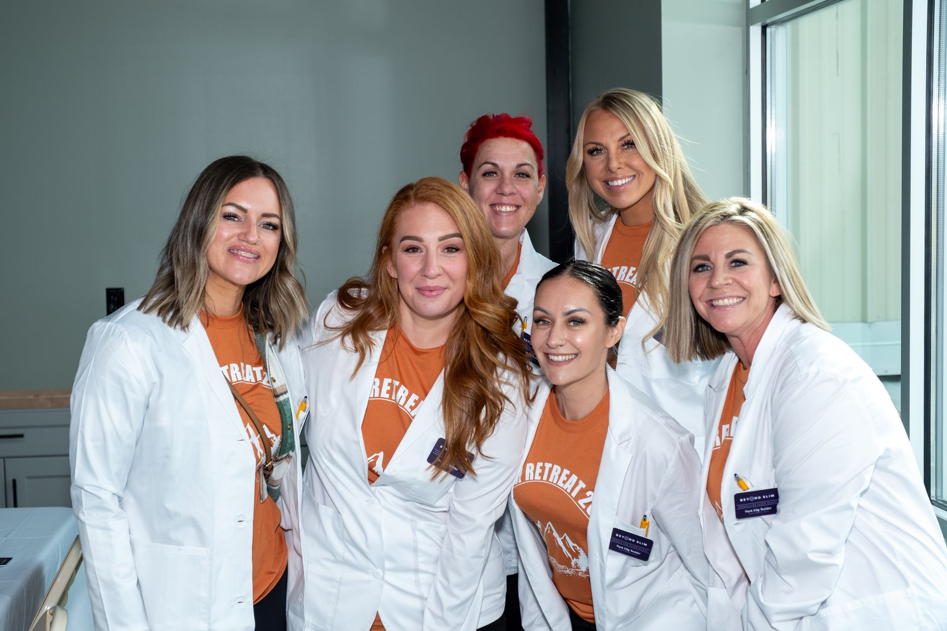 Six individuals wearing white lab coats over orange t-shirts stand together smiling in an office setting.