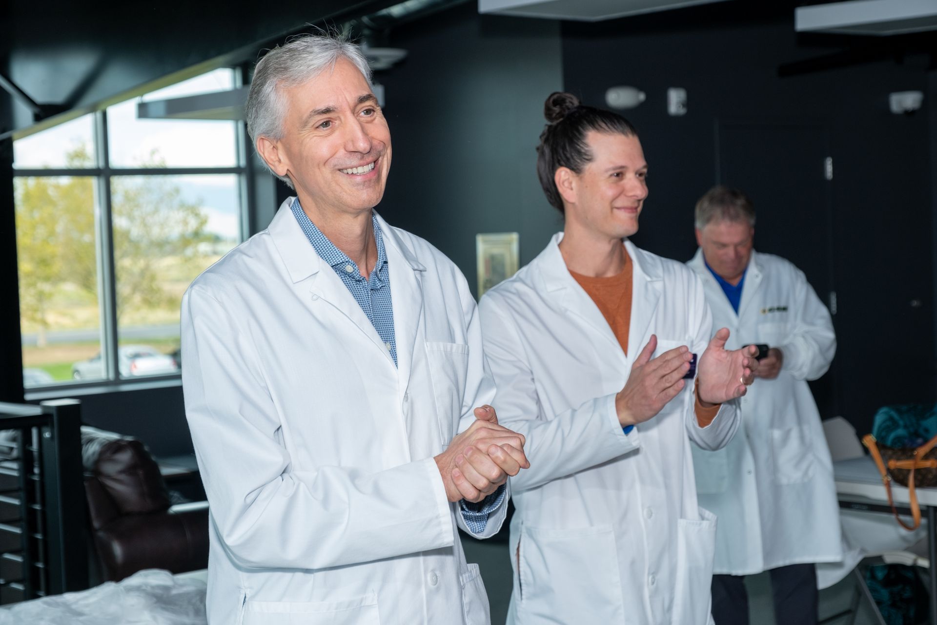 Three people wearing white lab coats stand indoors, smiling and clapping, while facing toward their right.