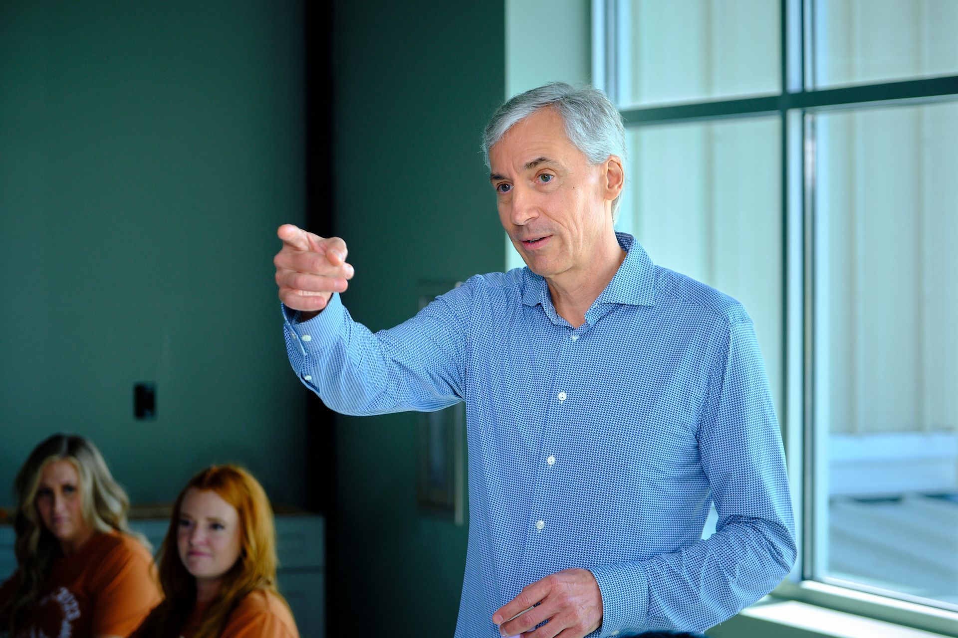 A person in a blue checkered shirt points toward an audience in a room with teal walls and a large window.