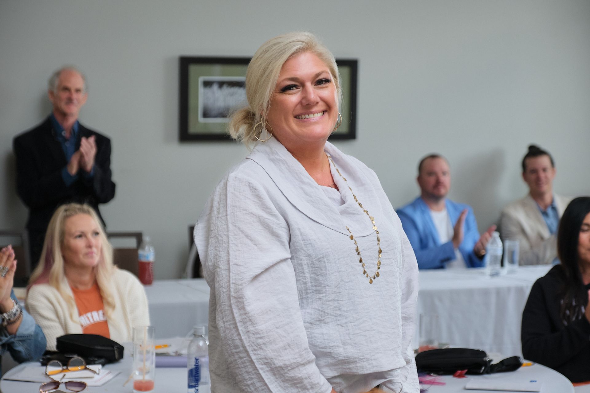 A smiling woman in a light-colored top stands in a room while others sit at tables and applaud in the background.
